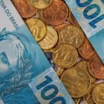 Cédulas de 100 reais dispostas diagonalmente sobre uma superfície coberta por diversas moedas brasileiras de diferentes valores, incluindo 1 real, 50 centavos e 5 centavos. A imagem mostra detalhes das notas e moedas em tons de azul, dourado e cobre, simbolizando finanças, economia e dinheiro no Brasil. Forneça seus comentários sobre o BizChat