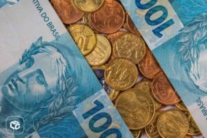 Cédulas de 100 reais dispostas diagonalmente sobre uma superfície coberta por diversas moedas brasileiras de diferentes valores, incluindo 1 real, 50 centavos e 5 centavos. A imagem mostra detalhes das notas e moedas em tons de azul, dourado e cobre, simbolizando finanças, economia e dinheiro no Brasil. Forneça seus comentários sobre o BizChat